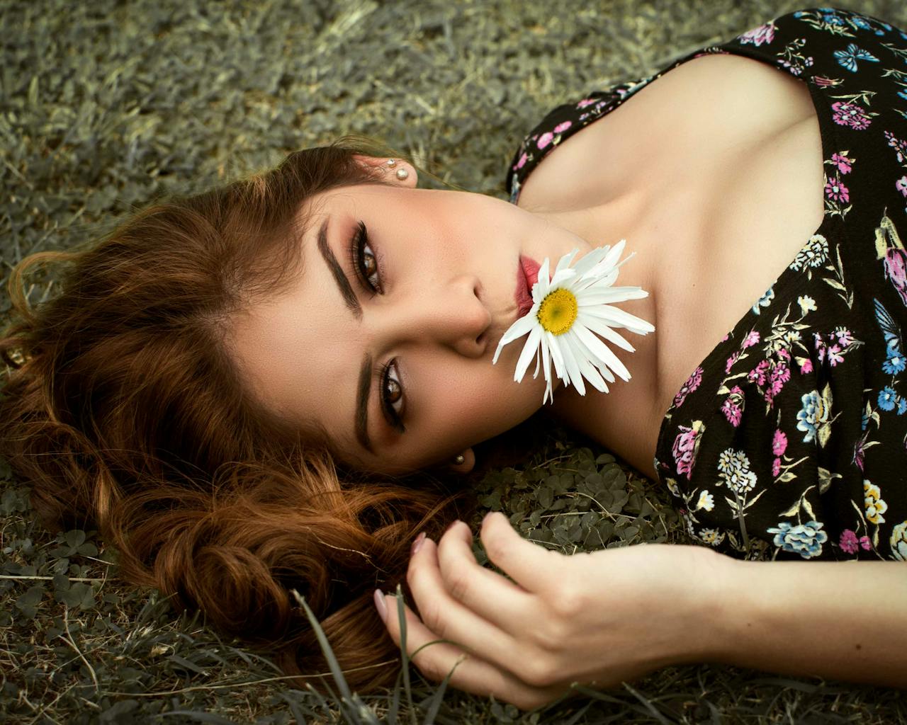 A young woman lies in grass holding a daisy, showcasing serene beauty and nature's charm.